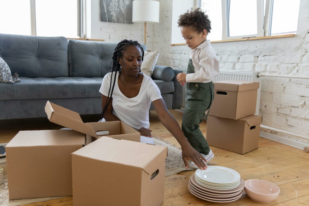 A mother and her son packing dishes into cardboard boxes in the living room.