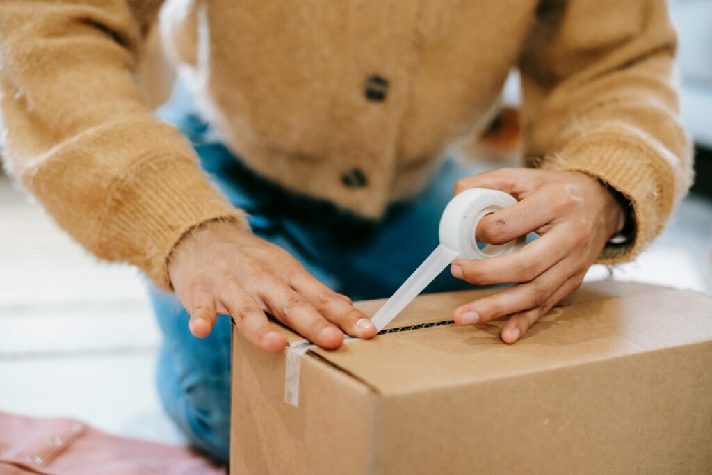 A woman uses tape to seal a cardboard box at home, preparing for shipping or moving.